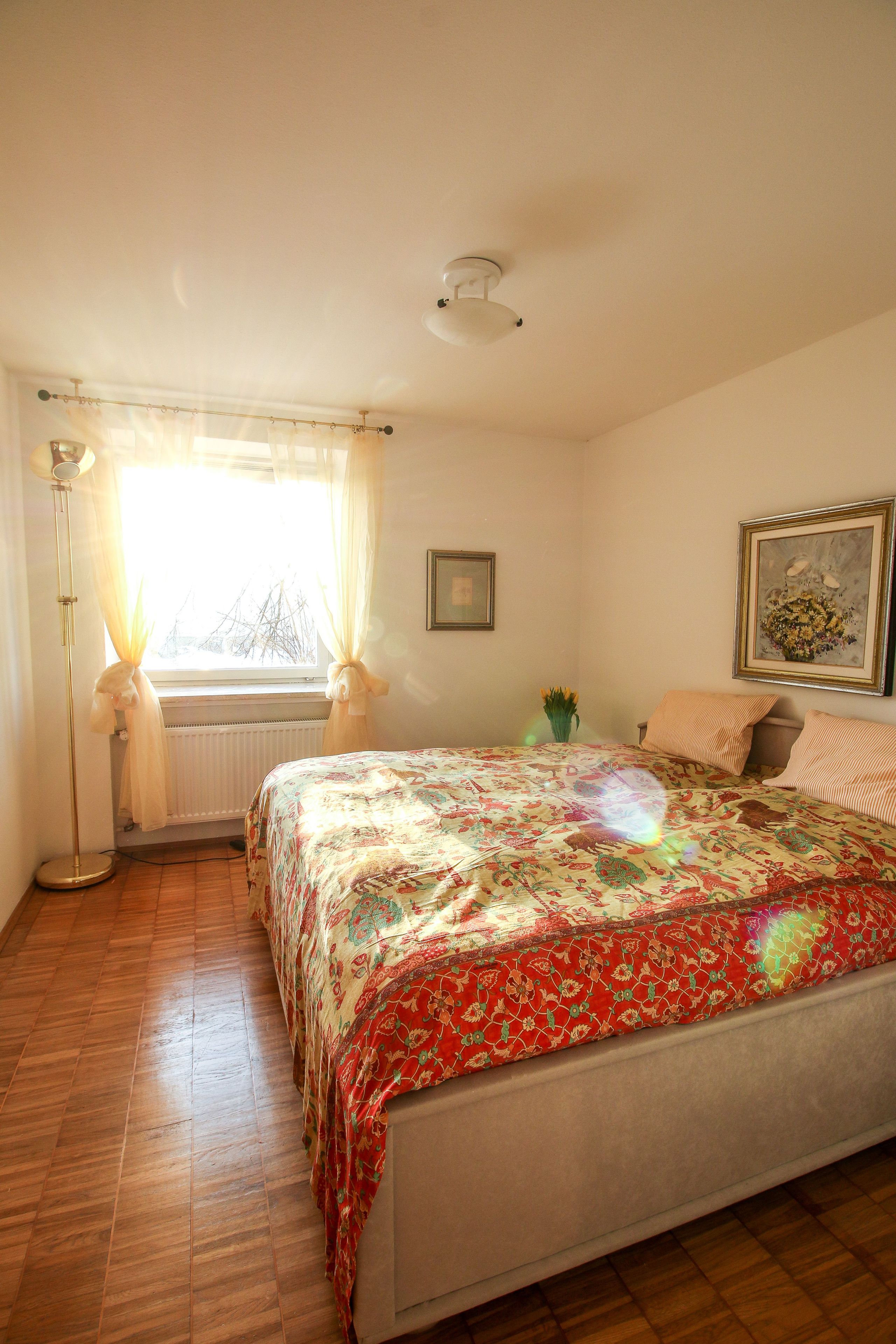 Een slaapkamer met een bed, een raam met gordijnen, een radiator, een vloerlamp en een schilderij aan de muur.