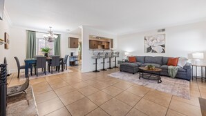 Ein geräumiges Wohnzimmer mit einer grauen Eckcouch, einem Couchtisch und einem Essbereich mit Tisch und Stühlen. Im Hintergrund ist eine Küche mit modernen Geräten und einer Bar erkennbar.
