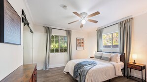 Ein Schlafzimmer mit Bett, Schreibtisch, Deckenventilator und einem Fenster mit Vorhängen.