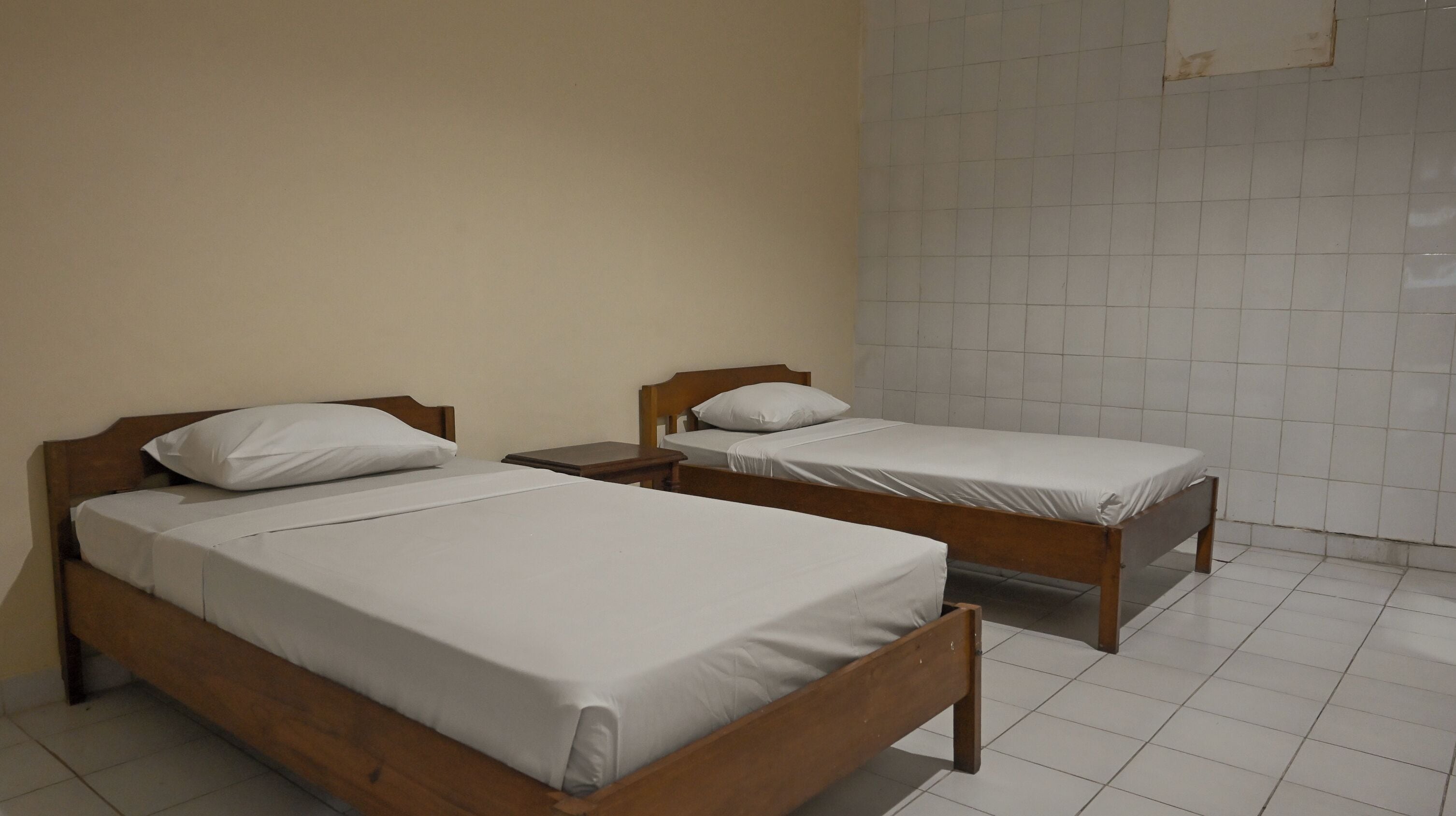 Two single beds with wooden frames and white bedding in a room with tiled flooring and plain walls.