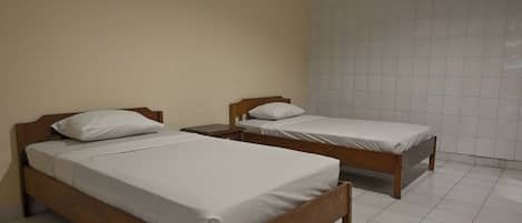 Two single beds with wooden frames and white bedding in a room with tiled flooring and plain walls.