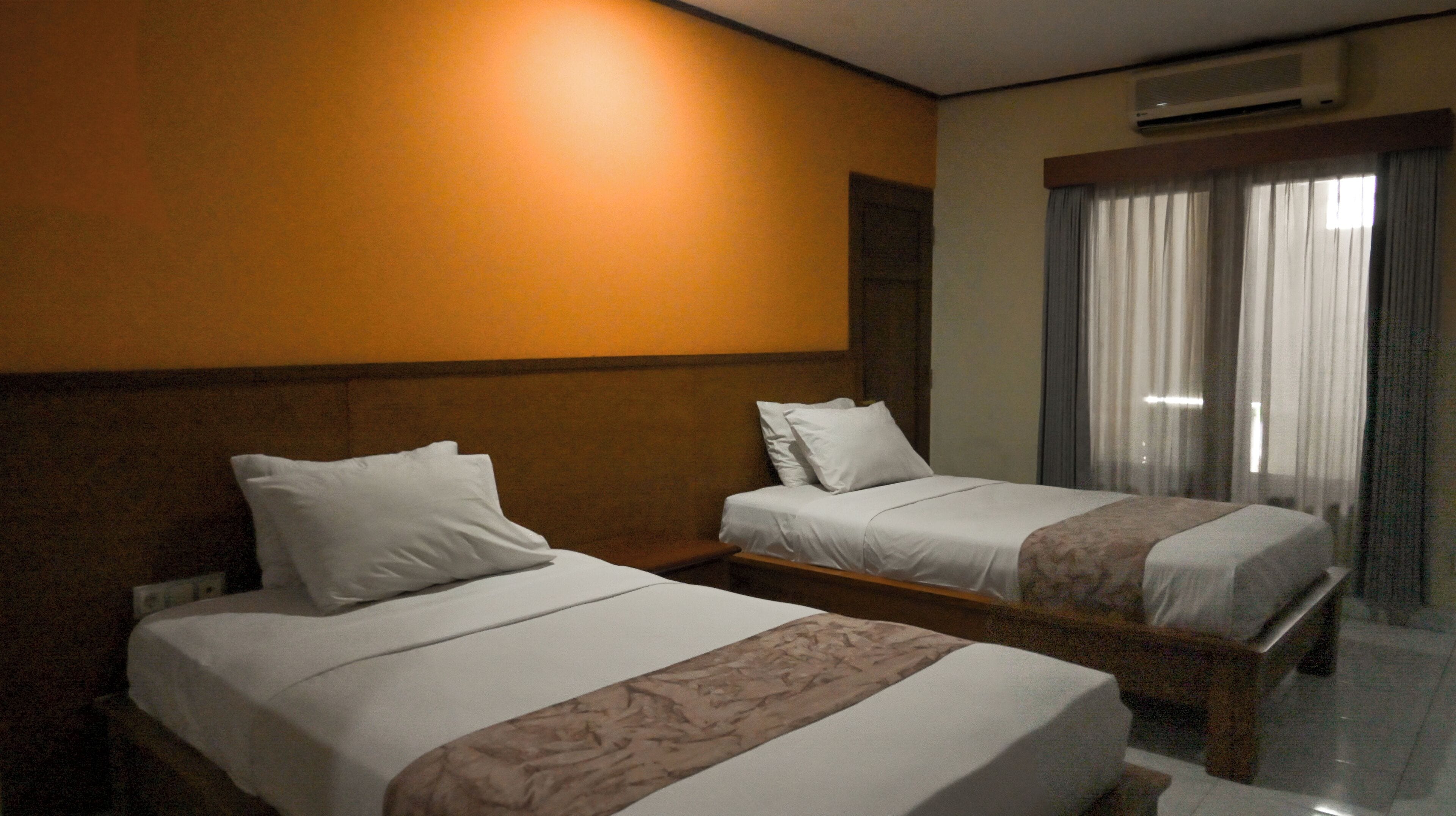 A hotel room with two beds, a wooden headboard, a large window with curtains, and an air conditioning unit on the wall.