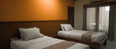 A hotel room with two beds, a wooden headboard, a large window with curtains, and an air conditioning unit on the wall.