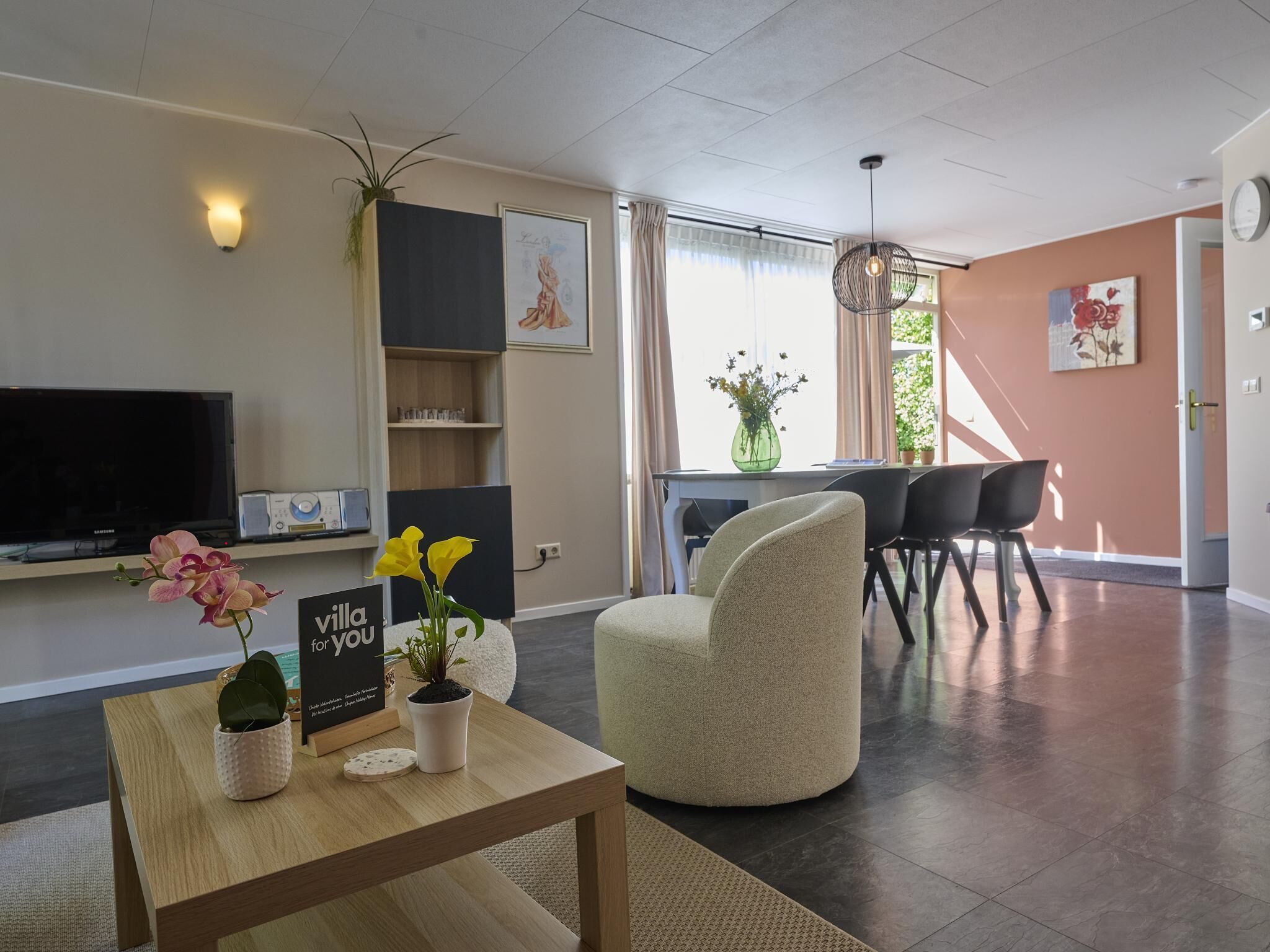 Una sala de estar moderna con un televisor de pantalla plana, una mesa de centro con plantas, un comedor con sillas y un ventanal con cortinas.