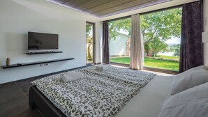 Quarto moderno com cama grande, televisão de tela plana na parede e vista para um jardim através de amplas janelas.