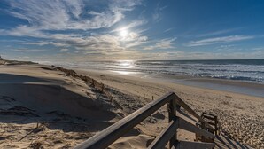 Een strand met een houten looppad dat naar het water leidt, een heldere lucht en de zon die op de oceaan schittert.