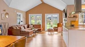 A spacious living area with a wooden ceiling, a sofa set, a dining table, and a kitchen area with a large window.