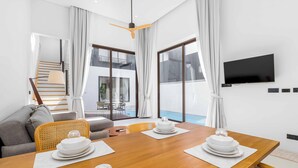 Ein modernes Hotelzimmer mit einem Holztisch, gedeckt für eine Mahlzeit, einer Couch, einem an der Wand befestigten Fernseher und einem Balkon mit Pool.