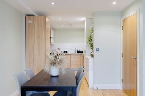 Un espace repas moderne avec une table et des chaises en bois, un vase contenant des plantes séchées et, au fond, une cuisine.