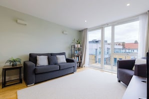 Ein modernes Wohnzimmer mit einem grauen Sofa, einem kleinen Beistelltisch mit Pflanze, einem Bücherregal und einem großen Fenster mit Blick auf die Stadt.