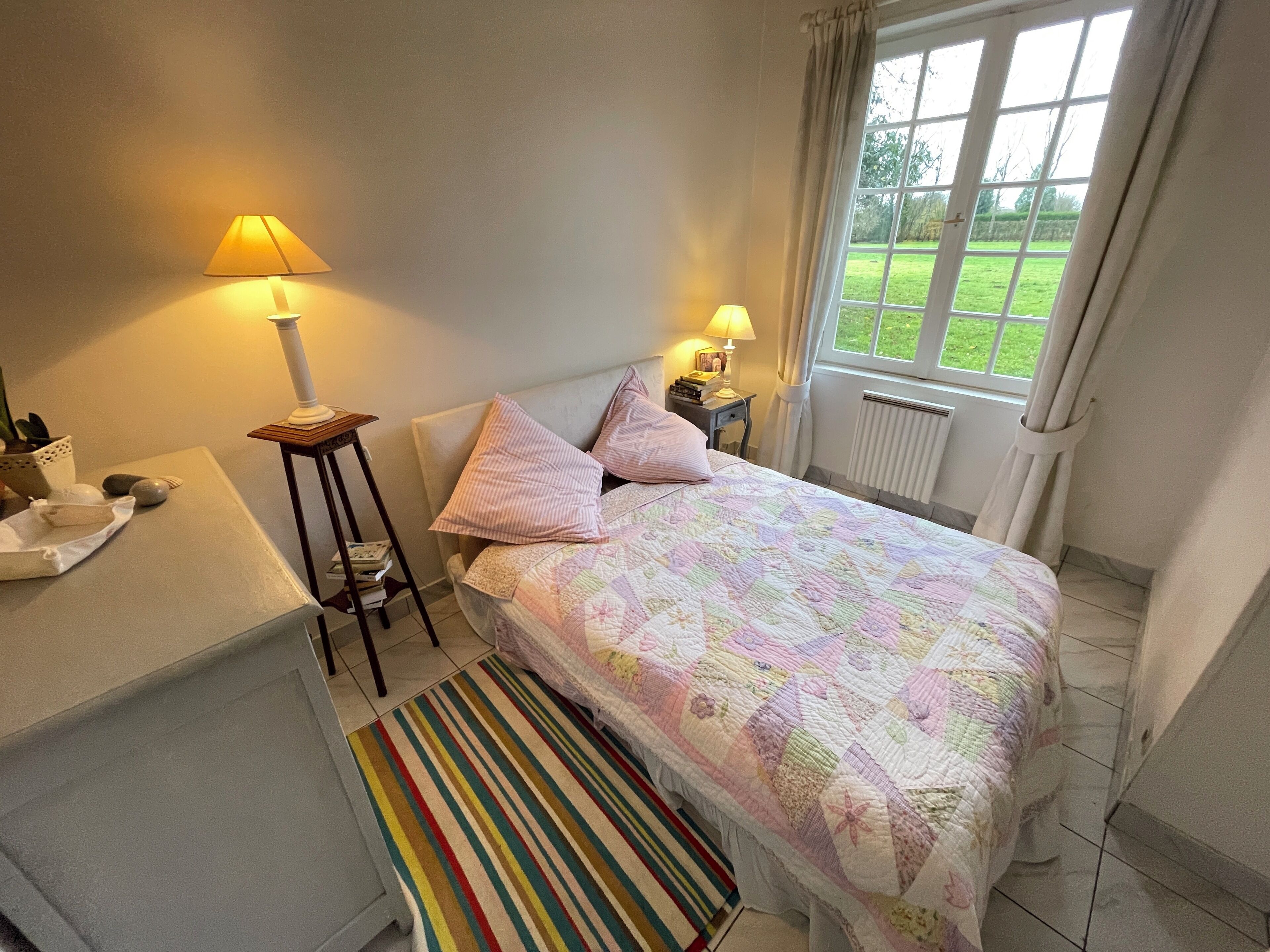 Une chambre à coucher avec un lit, deux lampes de chevet, une fenêtre avec des rideaux, un radiateur, une table de chevet avec une lampe, une petite plante et un tapis à rayures.