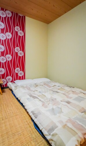 A small, single-bed room with a wooden floor, a plain wall, and a red patterned curtain. - meisei ebiya (Osaka)