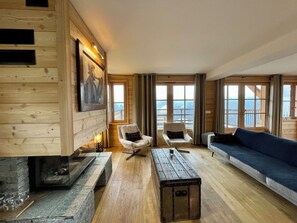 A cozy cabin-style room with a stone fireplace, wooden walls, and large windows offering a view of the outdoors. - Les Planches de Villard 5* Chalet – Terrace, sauna – Alpe d’Huez area (Villard-Reculas)