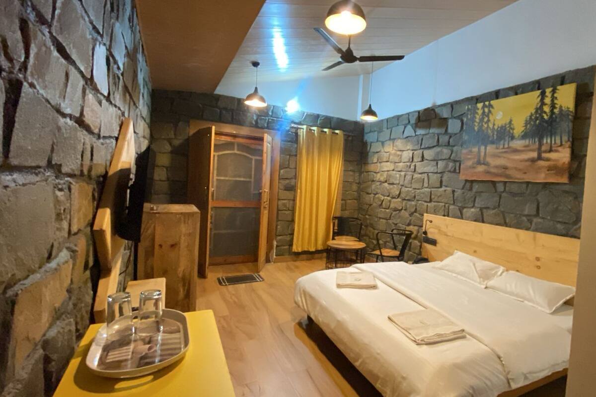 Um quarto com uma cama de madeira, uma mesinha com copos, uma cadeira, um guarda-roupa e uma parede de pedra.