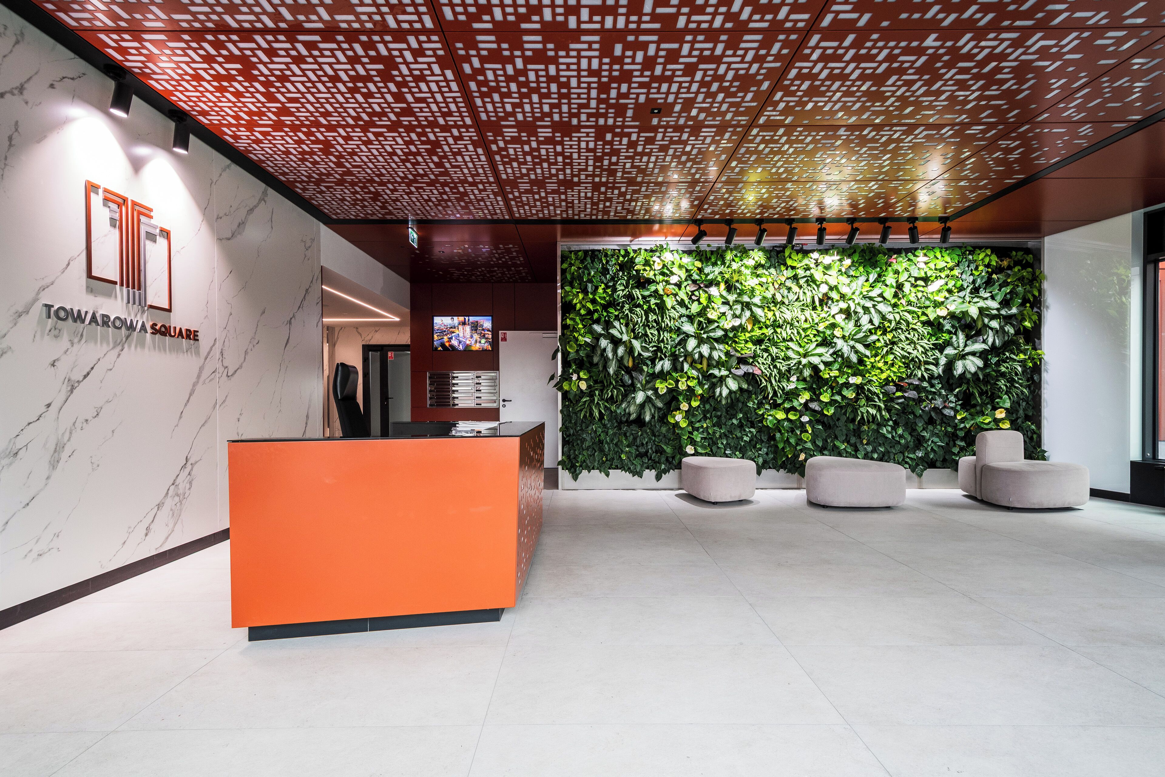 A modern reception area with a marble wall, an orange reception desk, a green vertical garden, and a ceiling with a geometric pattern.