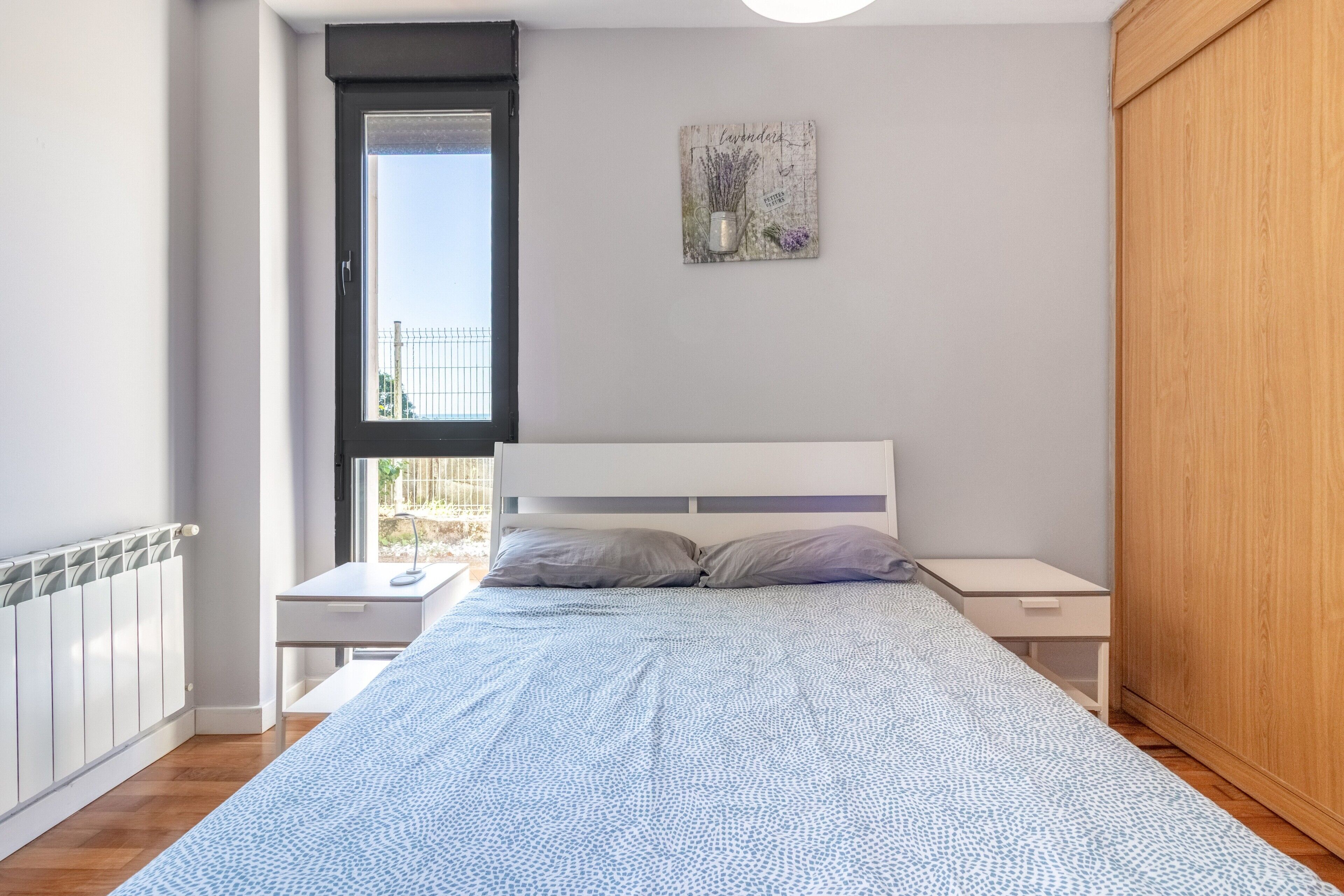 A bedroom with a bed, bedside tables, a window, a radiator, and a wooden wardrobe.