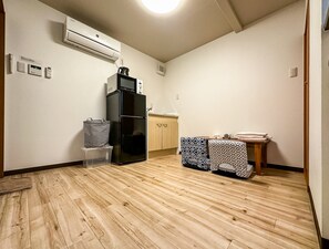 A compact room with a built-in air conditioner, a black refrigerator, a wooden cabinet, and patterned cushions on a small table. - meisei ebiyaminami (Osaka)