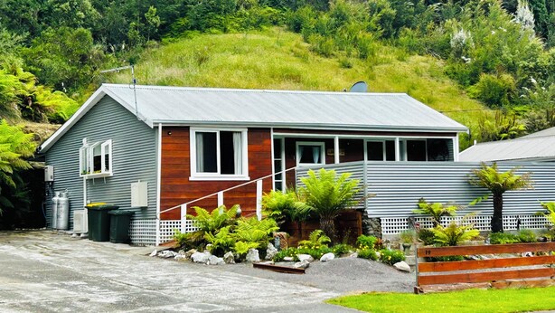 A single-story house with a sloped roof, a front porch, and a small garden area with various plants and a bench. - Cosy retro renovated cottage with mountain setting out back (Queenstown)