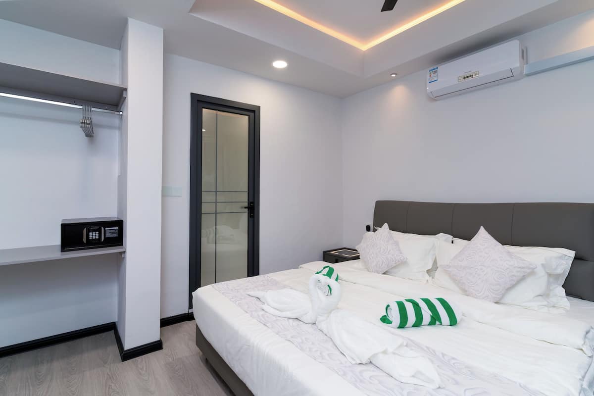 Une chambre d’hôtel moderne avec un lit bien fait, une table de chevet, un climatiseur fixé au mur et une porte en verre donnant sur la salle de bain.