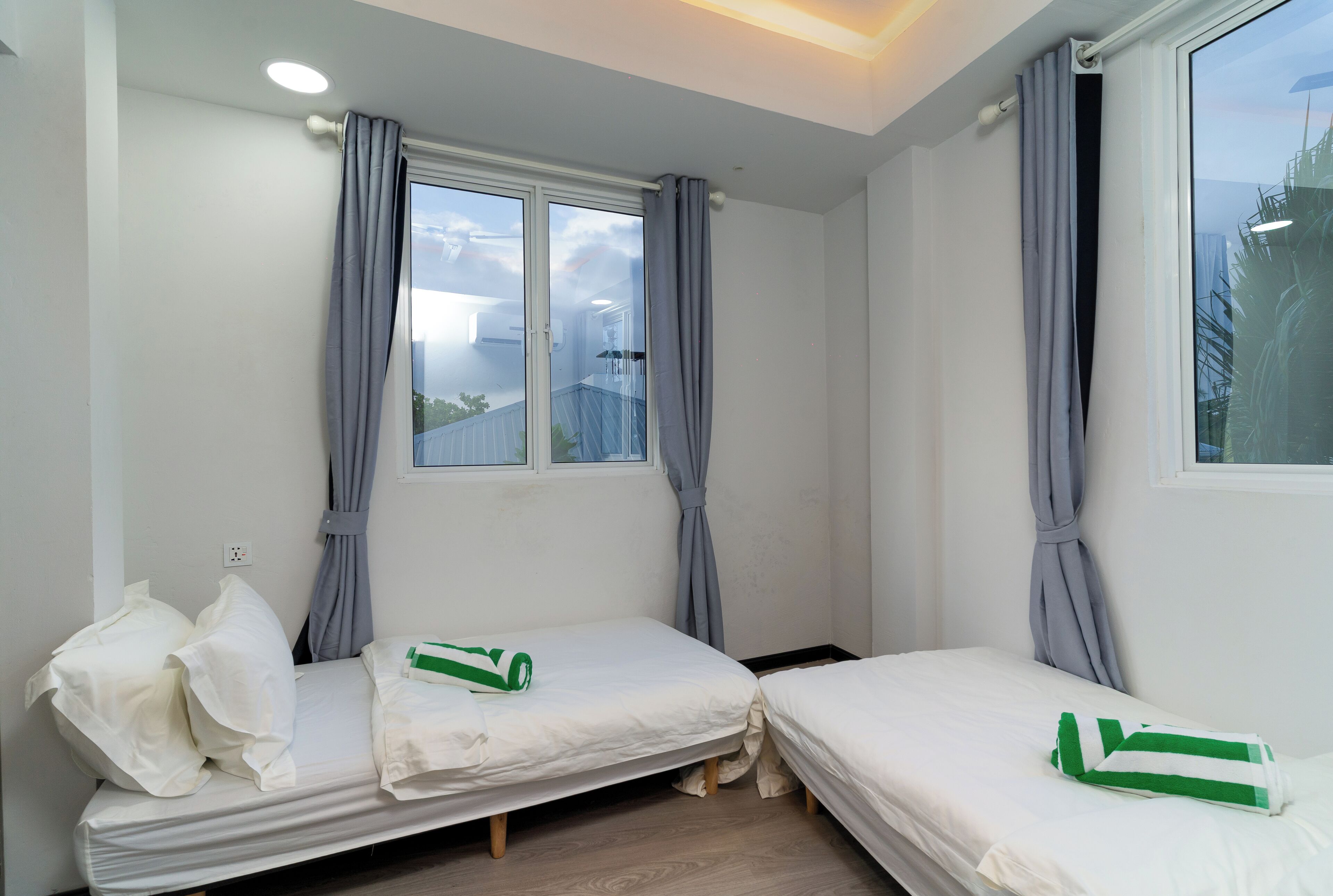 A small, clean room with two single beds, a window with curtains, and a view of the outside.
