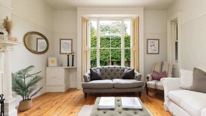 A living room with a grey sofa, a white armchair, a coffee table with books, a large window with a view of greenery, and a fireplace with a mantelpiece. - House & Annexe, Sleeps 18, Heated Pool (Sidlesham)