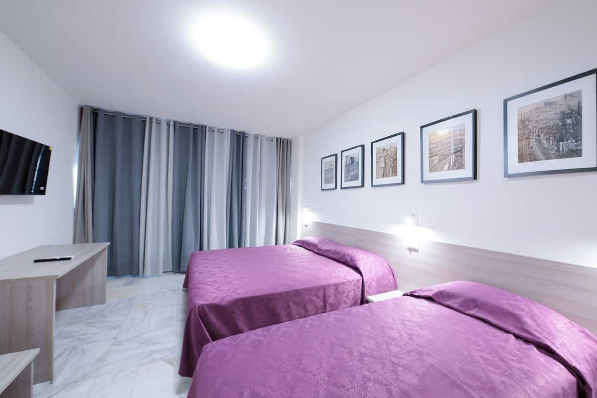 A hotel room with two beds, a desk, a TV, and framed artwork on the wall.