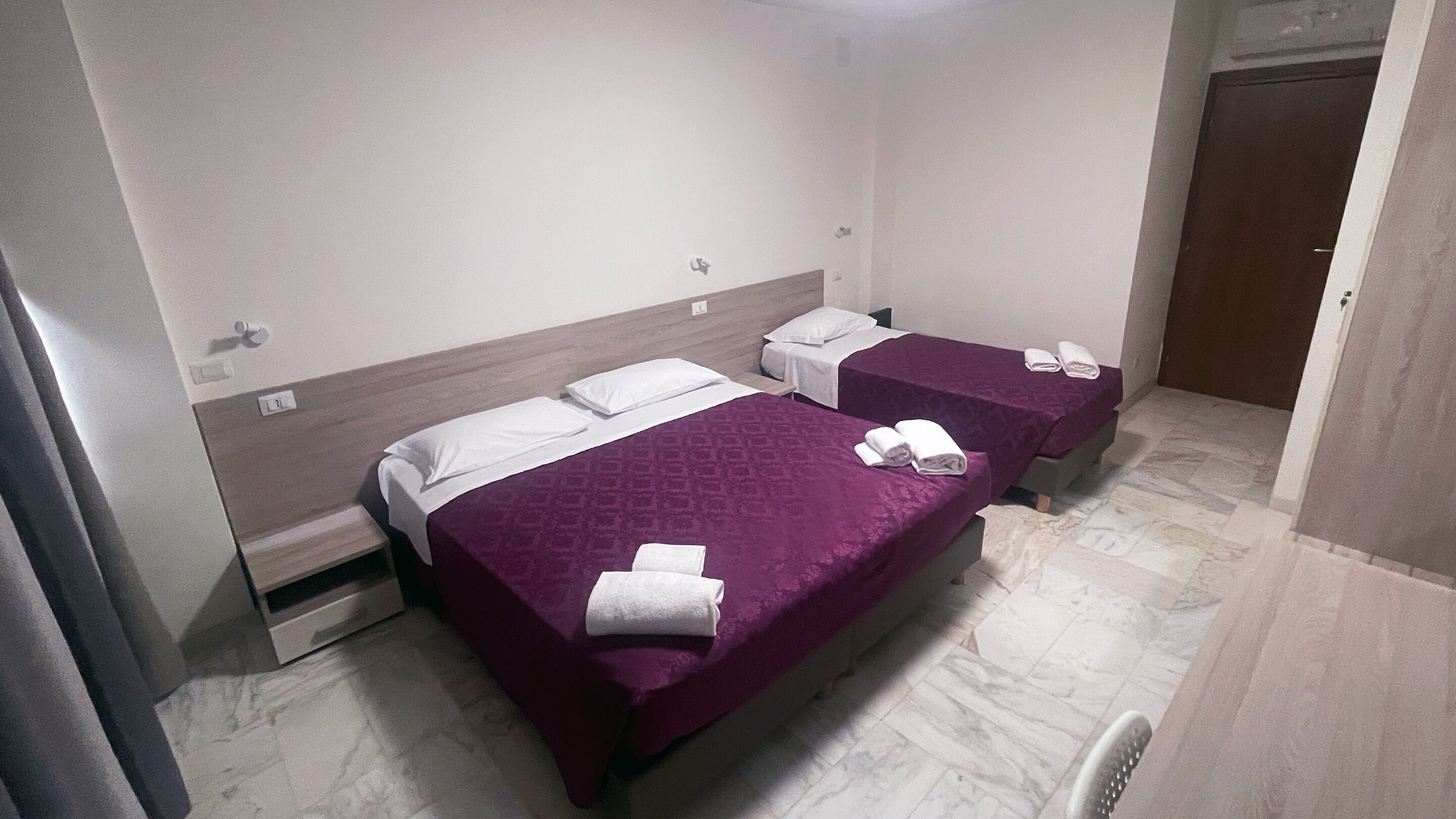 A hotel room with two beds, each with a purple bedspread and white pillows. There are towels neatly folded on each bed.