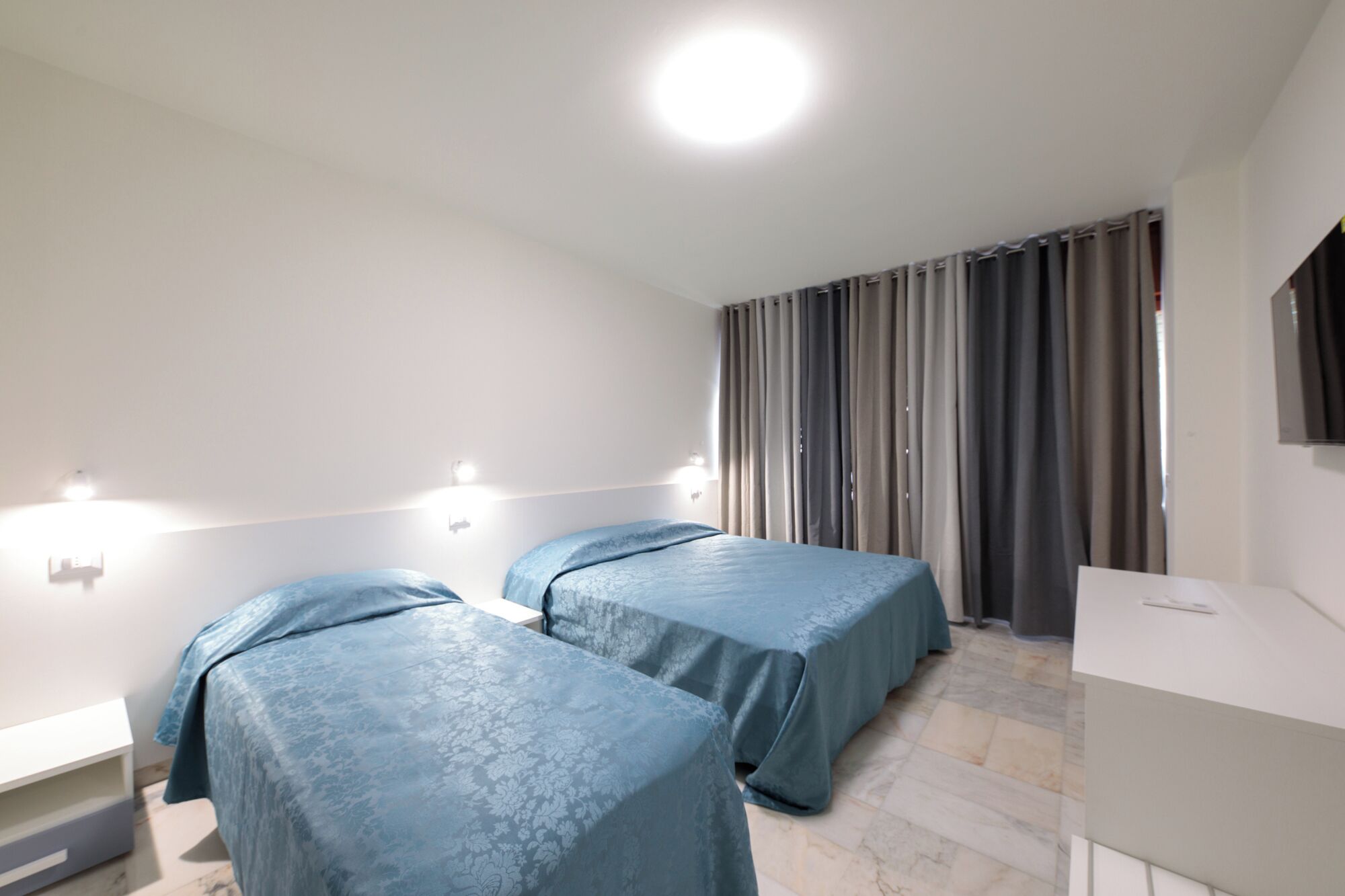 A hotel room with two beds, a bedside table, a mirror, and a window with curtains.