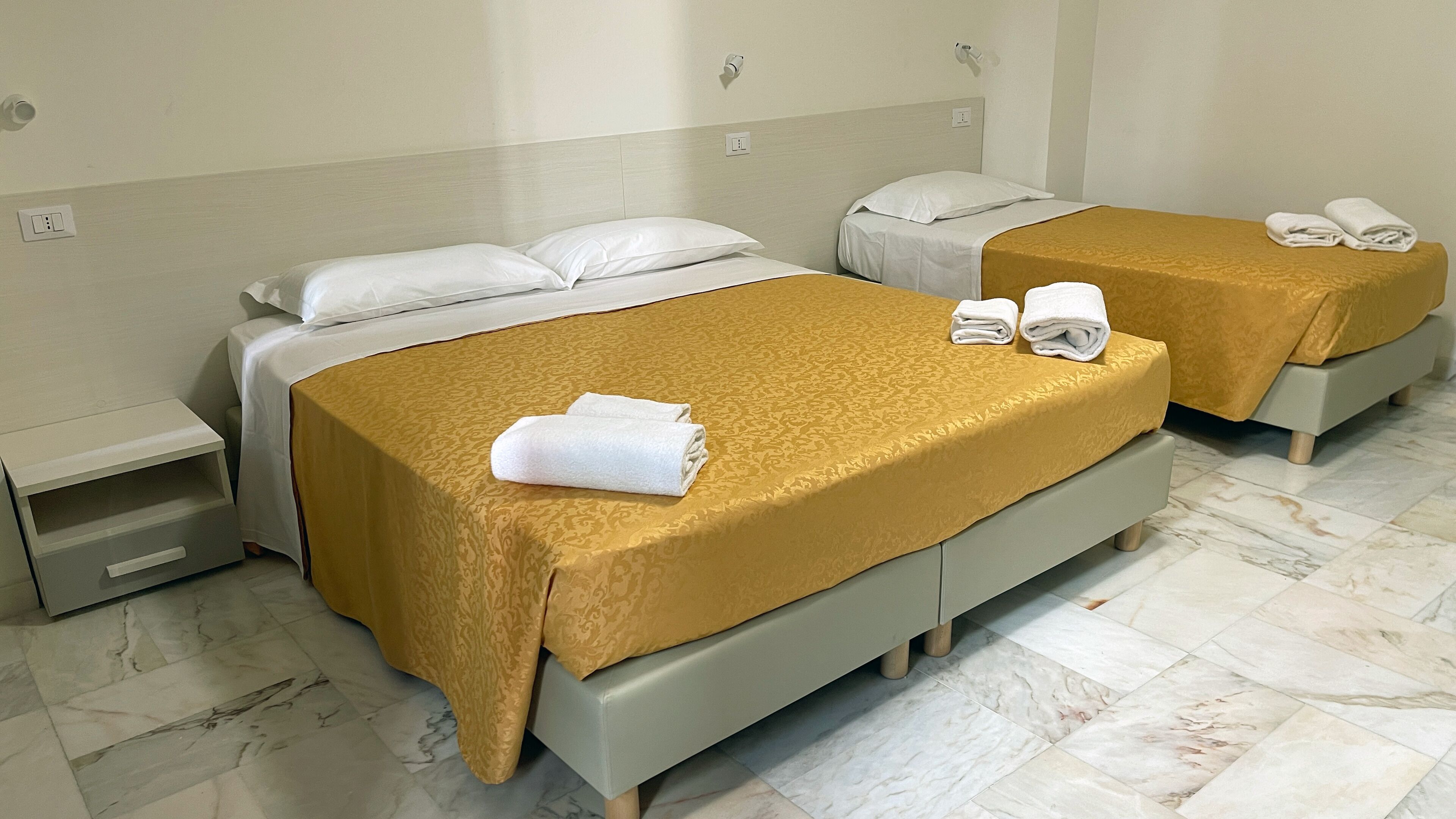 Une chambre d’hôtel avec deux lits, chacun agrémenté d’un oreiller blanc et d’une couvre-lit dorée. Une petite table de chevet avec un tiroir se trouve sur le côté gauche de la pièce.