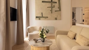 A modern living room with a beige sofa, a round coffee table with a vase of flowers, a flat-screen TV mounted on the wall, and a minimalist abstract painting. - Elegant apartment with AC - Le Marais (Paris)