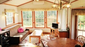 A cozy living room with a sofa, a wooden coffee table, a dining table with chairs, a television, and large windows with curtains. - Cottage Paradiso (Onna)
