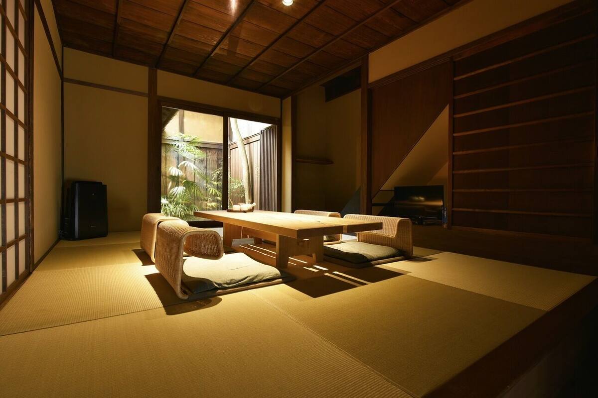Een kamer met tatami matten, een lage tafel, kussens, een schuifdeur en uitzicht op groen buiten.