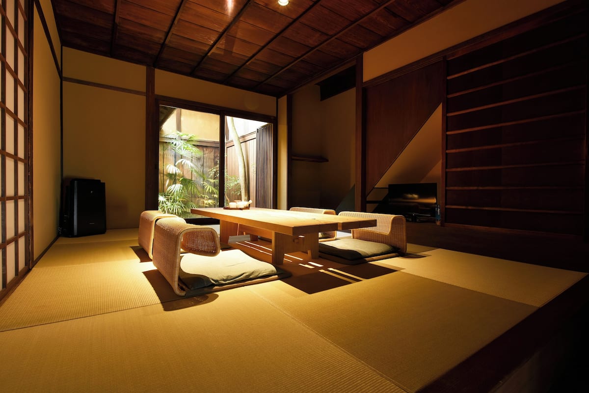 Een kamer met tatami matten, een lage houten tafel en kussens, een schuifdeur en uitzicht op de omgeving.