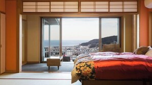 A hotel room with tatami flooring, a bed with a floral bedspread, and a view of a coastal town. - Bettei Sora no ue (Ito)