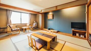 Een traditionele Japanse kamer met tatami vloerbedekking, een lage tafel, stoelen, een televisie en een plank met spullen.