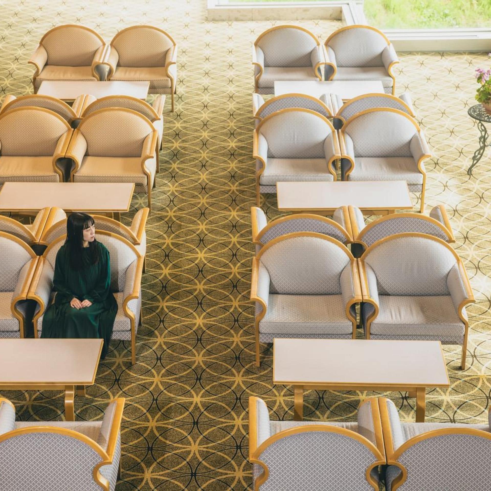 Habitación de hotel con alfombra de estampado geométrico, una persona sentada en una mesa y una ventana con vistas.