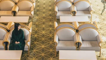 Habitación de hotel con alfombra de estampado geométrico, una persona sentada en una mesa y una ventana con vistas.