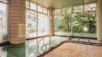 Een traditioneel Japans onsen met een betegelde vloer, een houten plafond en grote ramen met uitzicht op het omliggende bos.