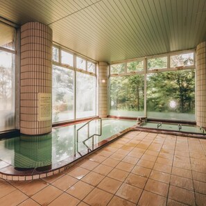 Un onsen japonés tradicional con suelo de baldosas, techo de madera y grandes ventanales que dan a un bosque circundante.