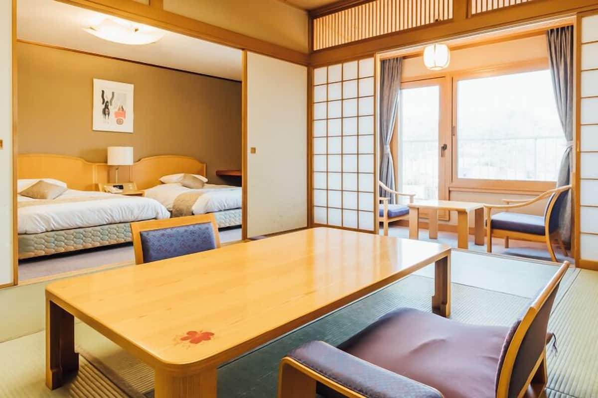 A traditional Japanese-style room with tatami flooring, a low wooden table, a cushioned bench, a bed with a headboard, and a sliding shoji screen.