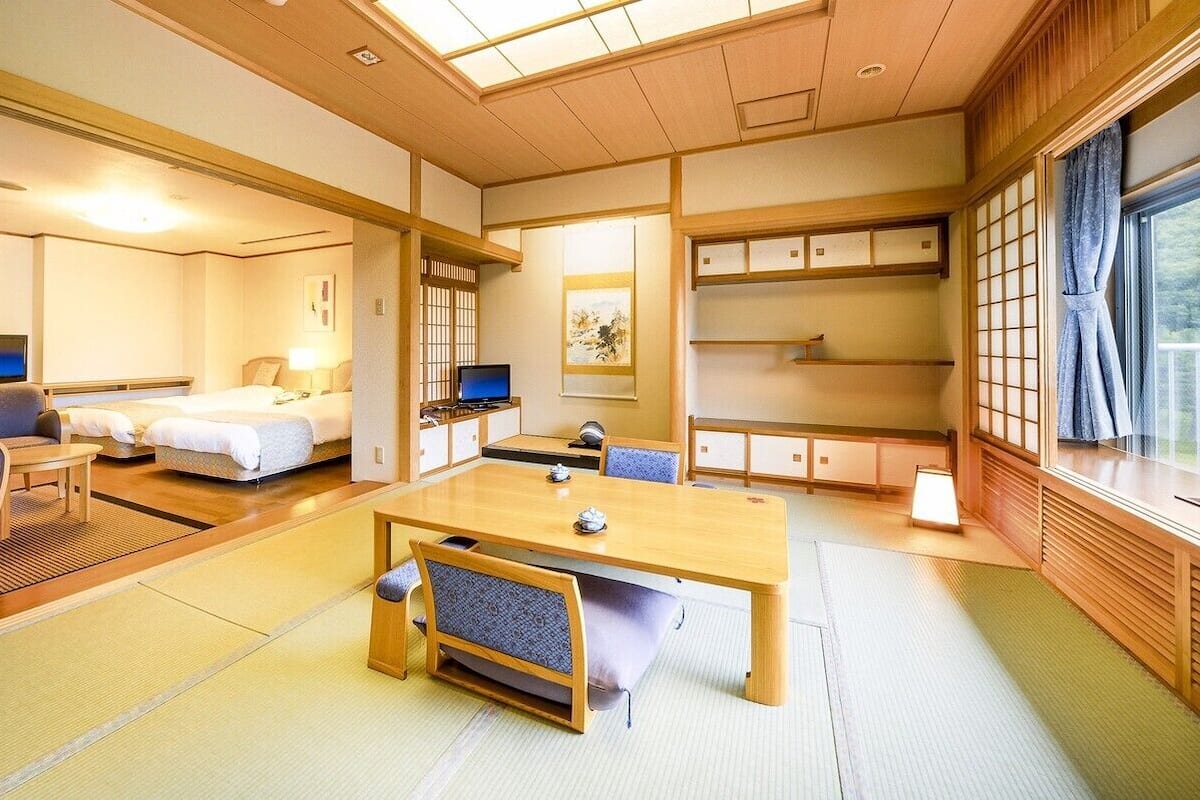 A traditional Japanese-style hotel room with tatami flooring, a wooden table, a sofa, a TV, and a balcony with a view.