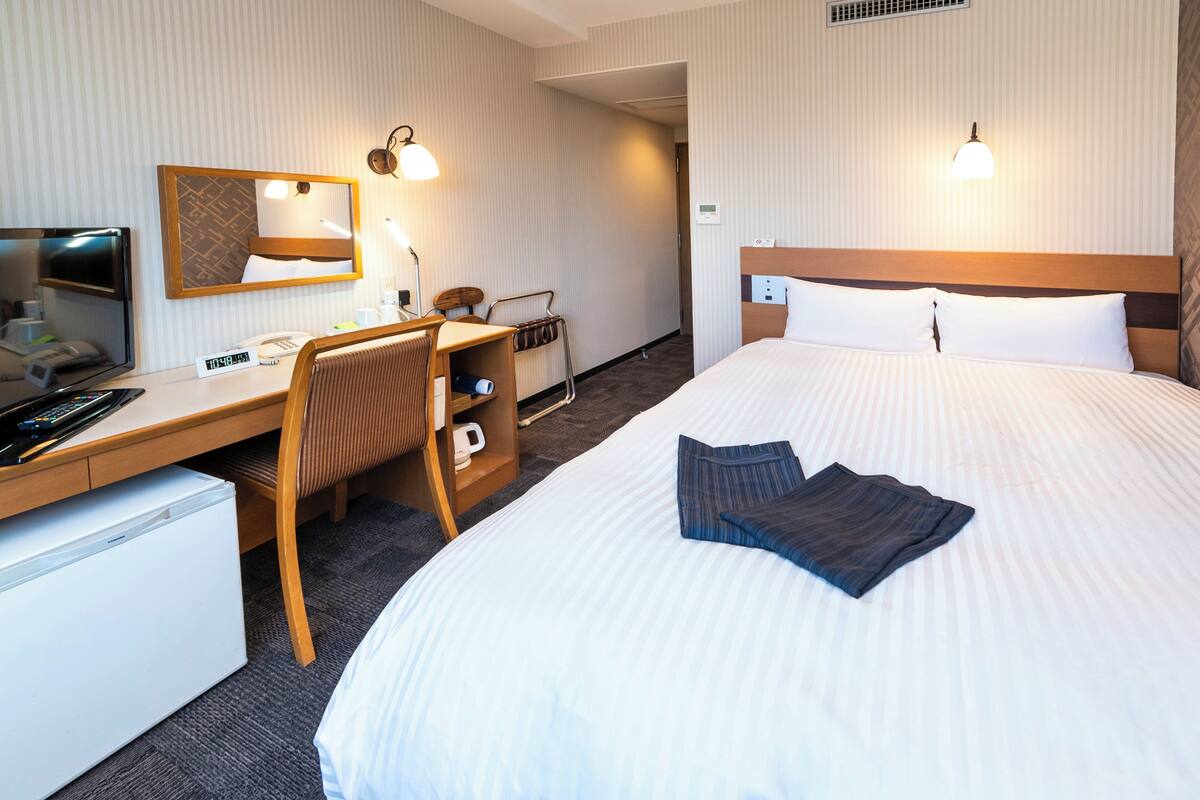 A hotel room with a bed, desk, chair, and a small refrigerator.