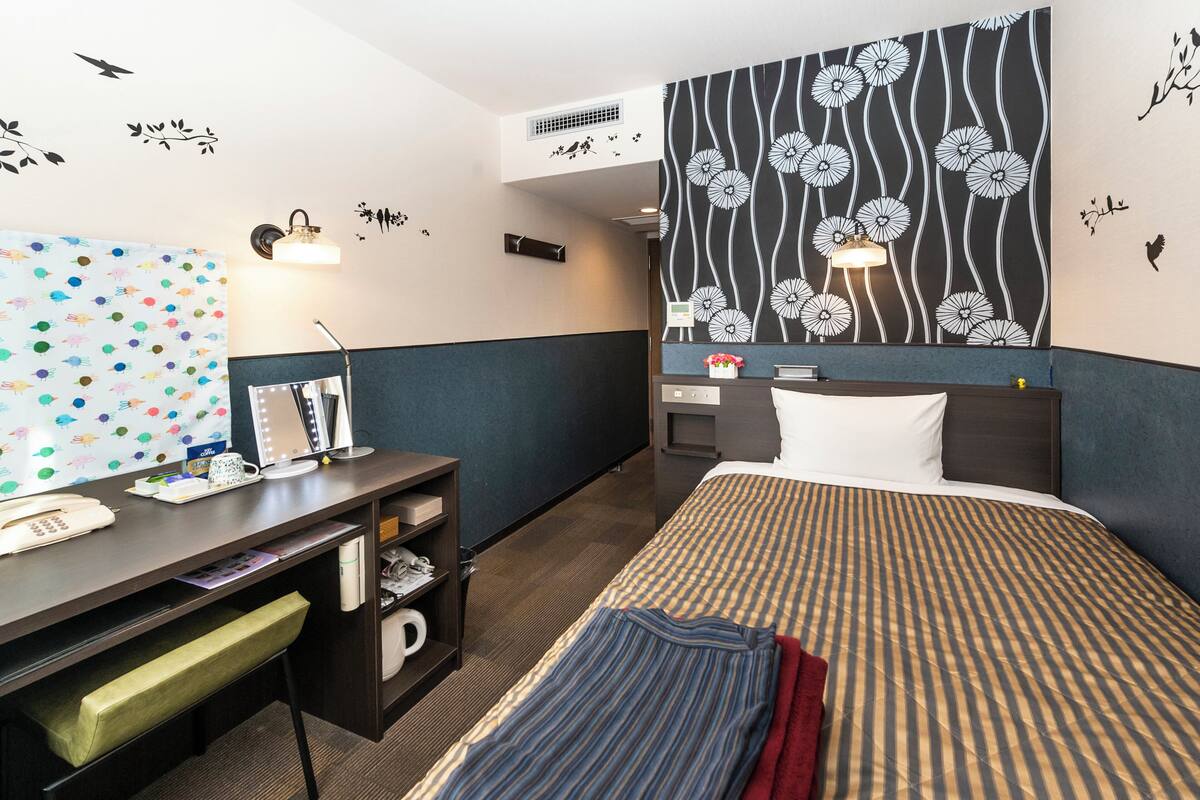 A hotel room with a bed, desk, chair, and a large wall decoration.