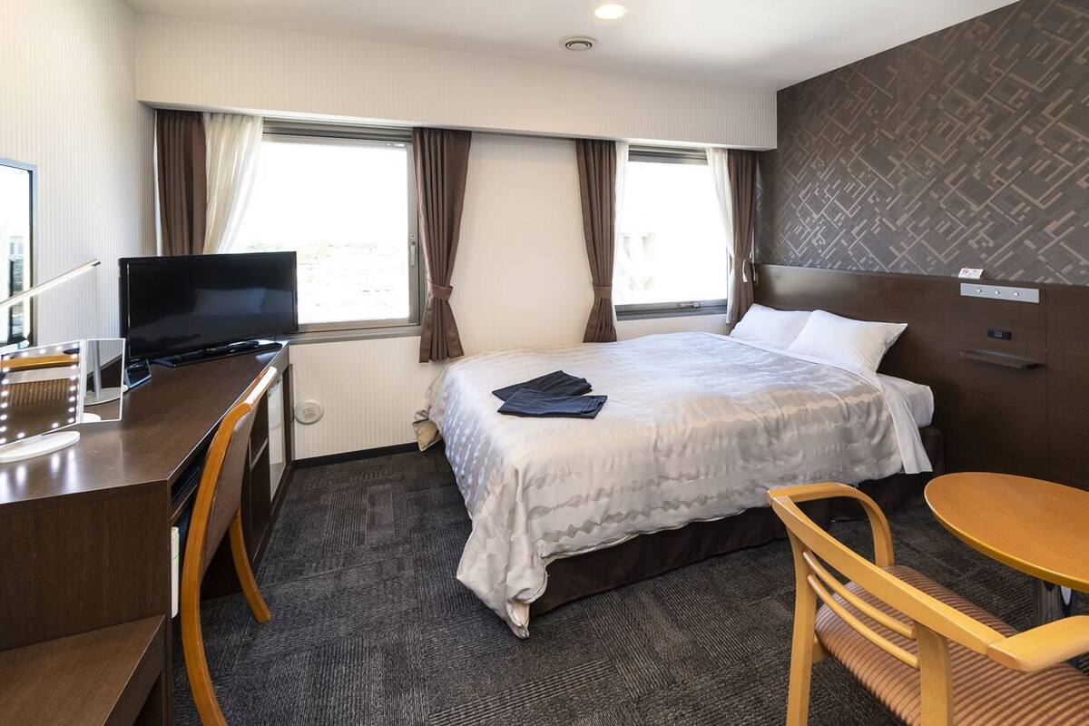 A hotel room with a bed, a desk with a TV, a chair, and a small table.