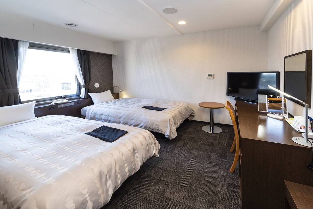 A hotel room with two beds, a TV, a desk with a chair, and a window with curtains.