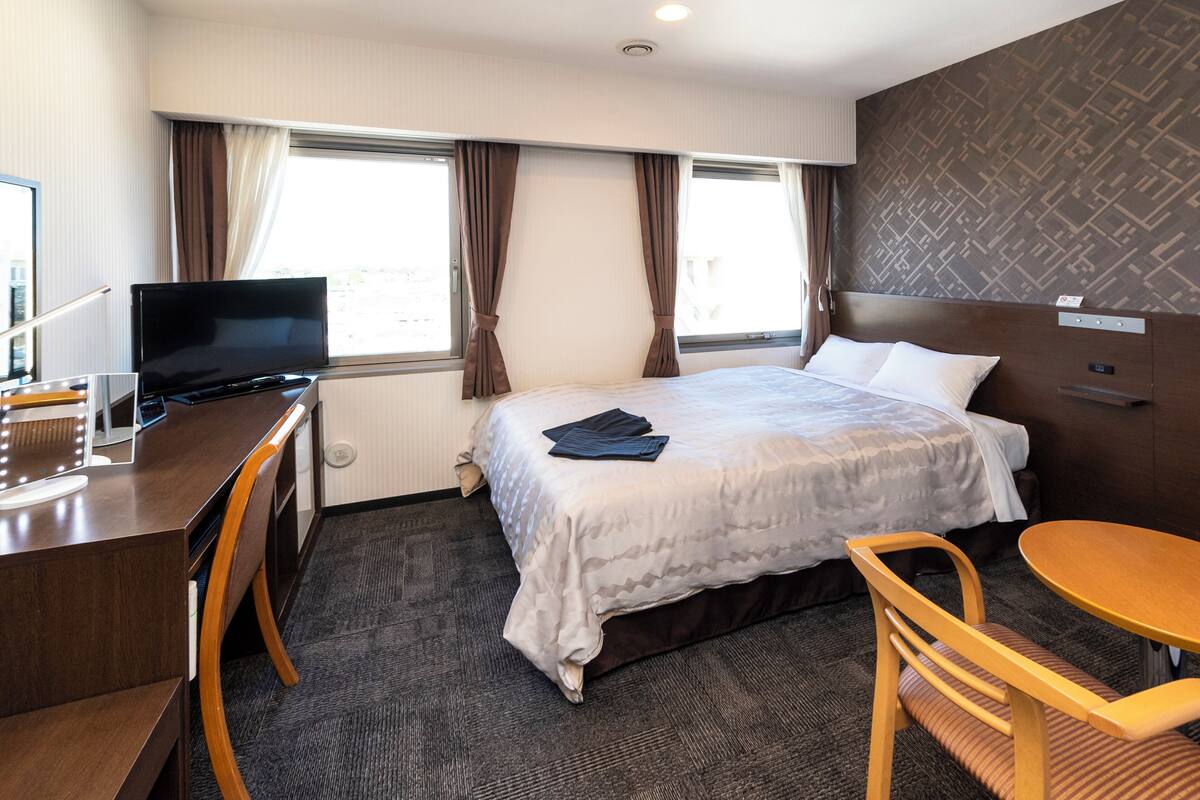A hotel room with a bed, a desk with a TV, a chair, and a small table.
