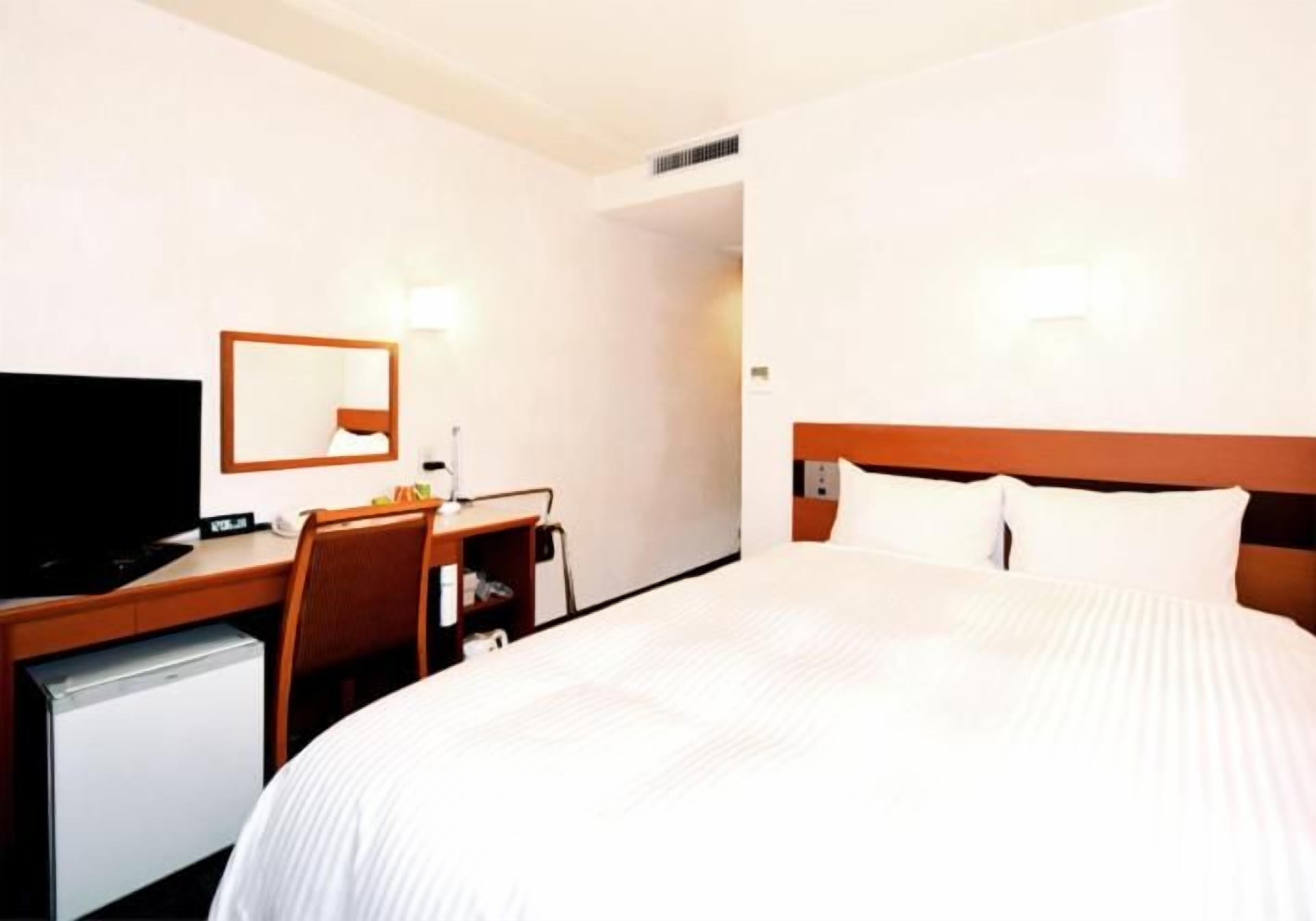 A hotel room with a bed, a desk with a chair, a television, and a small refrigerator.