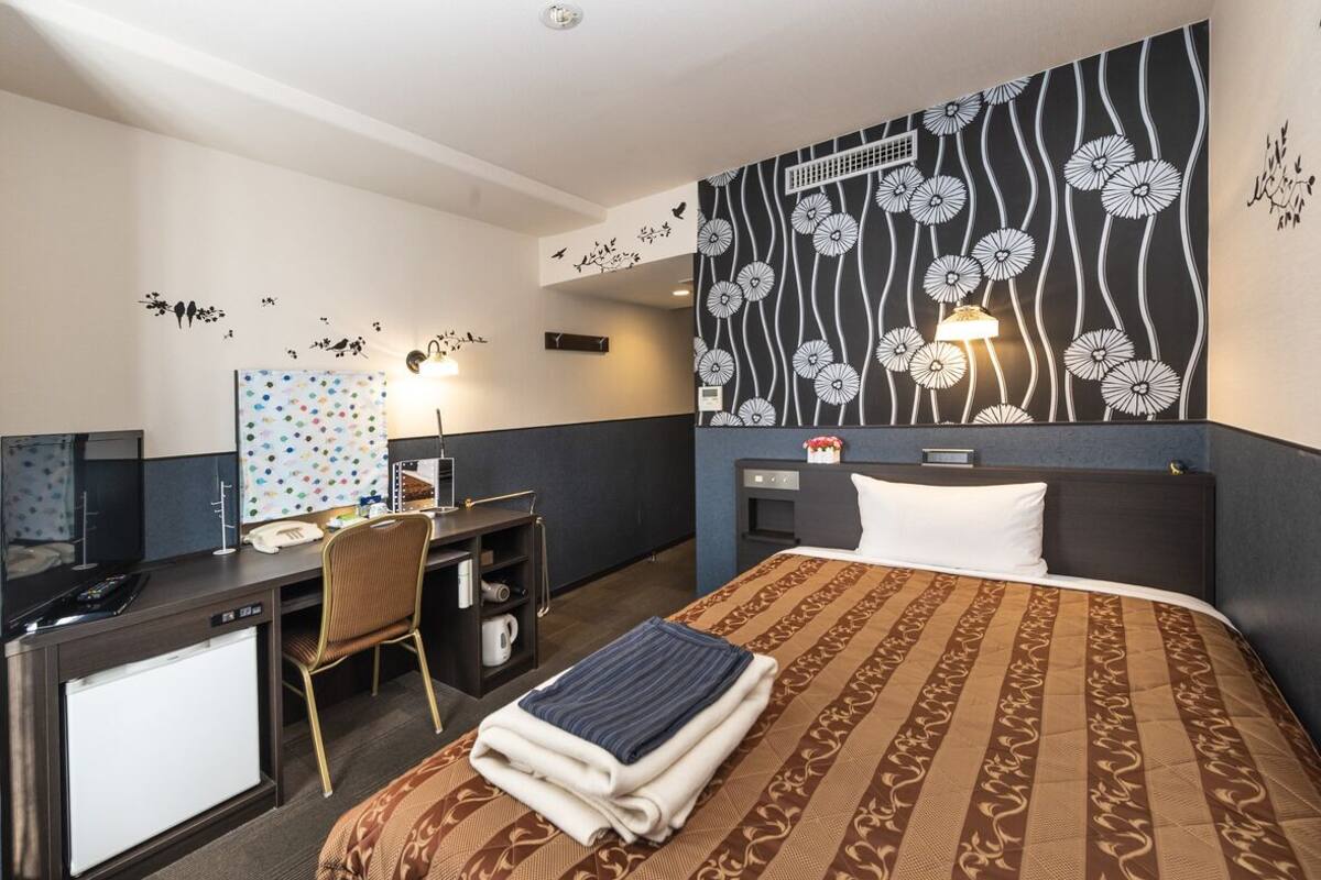 A hotel room with a bed, desk, chair, and a wall with a floral pattern.