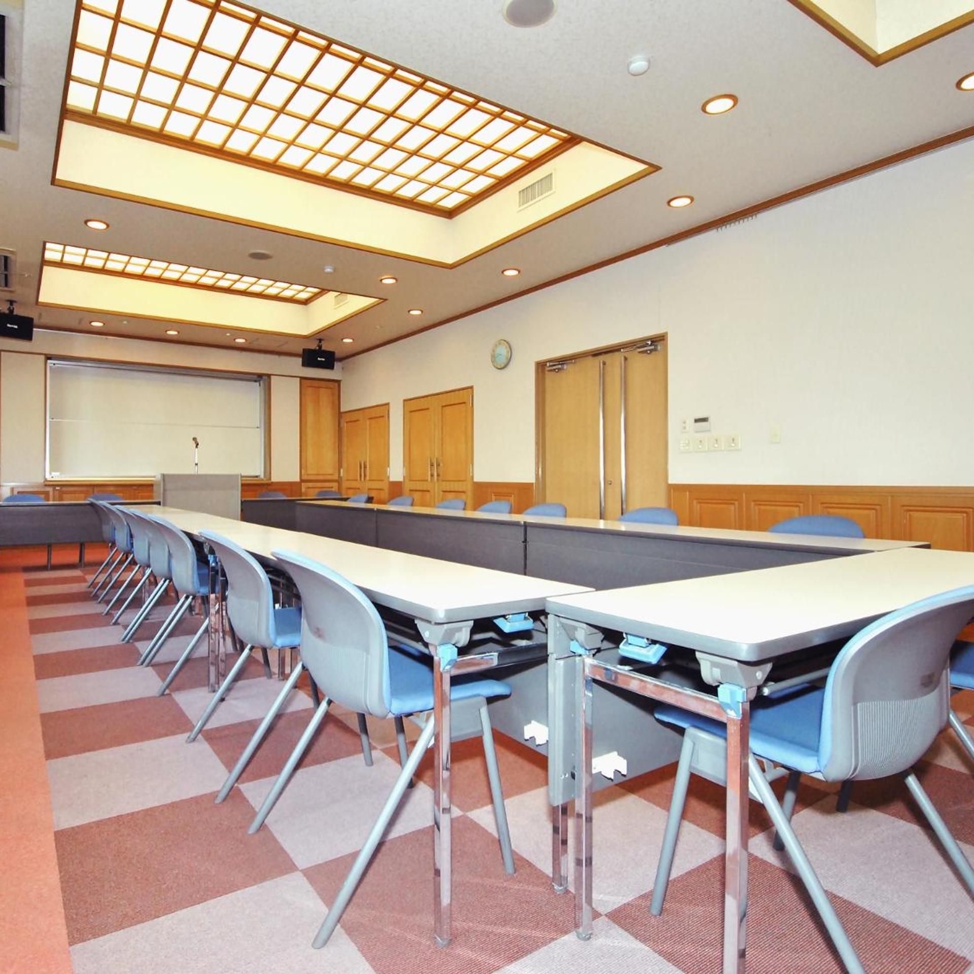 Sala de conferências com uma mesa comprida, cadeiras, tela de projetor e um relógio na parede.