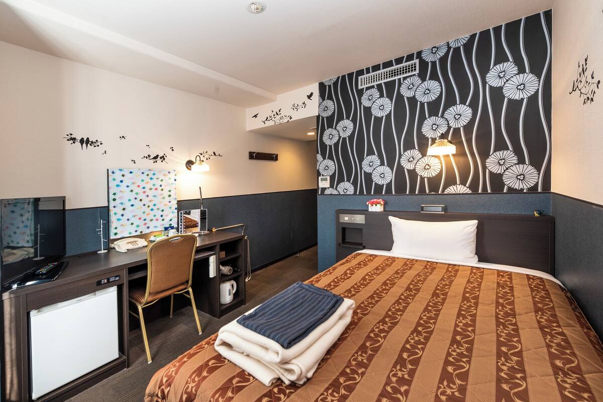 A hotel room with a bed, desk, chair, and a wall with floral patterns.